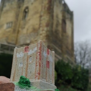 May include: A miniature stone castle model with red brick details, set on a wooden base with green foliage. The model is in front of a larger, weathered stone castle.