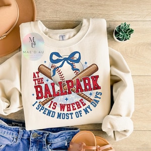 Op de afbeelding: Een crèmekleurige trui met een baseball-thema. De afbeelding toont een honkbal, twee gekruiste knuppels, een blauwe strik en de tekst "At the Ballpark is where I spend most of my days".