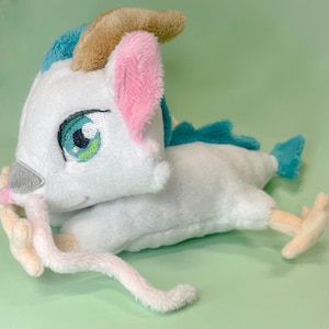May include: A white plush toy dragon with a light blue and brown horn, pink ears, and green eyes. The dragon is lying on its side with its tail curled around its body.