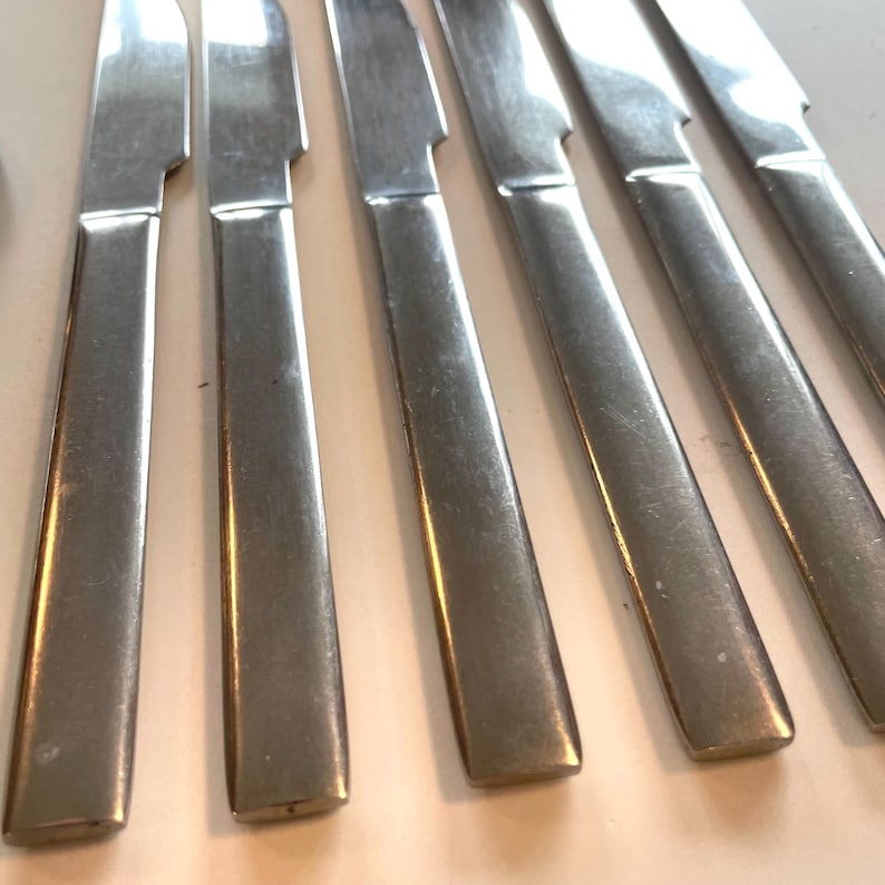 May include: A set of six stainless steel dinner knives. The knives have a simple, modern design with a rectangular handle and a serrated blade. The blades are angled slightly, and the handles are smooth and polished.