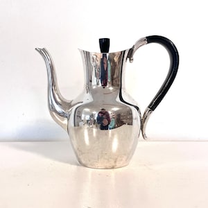 May include: A silver coffee pot with a black handle and lid. The pot has a curved spout and a rounded body. The handle is black and curved, and the lid is a small black cylinder. The pot reflects the surrounding environment.