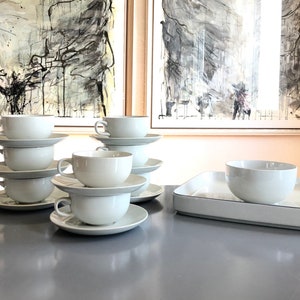 May include: A collection of white ceramic teacups and saucers, stacked in sets. A rectangular serving tray and a bowl are also present. The items are arranged on a gray surface, with abstract art in the background.