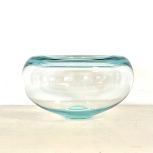 Puede incluir: Un cuenco de vidrio transparente con forma redondeada y borde azul claro. El cuenco tiene una abertura ancha y una base ligeramente aplanada. El vidrio es liso y transparente. El cuenco se utiliza probablemente con fines decorativos.