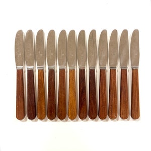 May include: A set of twelve butter knives with stainless steel blades and wooden handles. The handles vary in shades of brown, from light to dark. The knives are arranged in a row on a white surface.