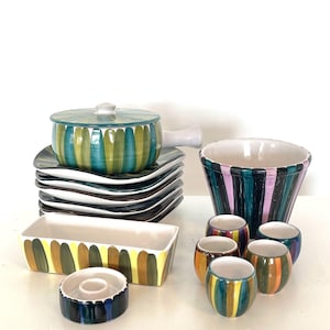 May include: A collection of colorful ceramic tableware with vertical striped patterns. The set includes a lidded pot, plates, a rectangular dish, a bowl, and small round cups. The color palette features shades of green, blue, yellow, and brown.