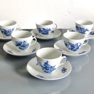 Könnte beinhalten: Set aus sechs weißen Porzellan-Teetassen und Untertassen. Jede Tasse und Untertasse hat ein blaues Blumendesign. Die Tassen haben ein strukturiertes Korbgeflechtmuster. Die Untertassen haben ein passendes Blumendesign.
