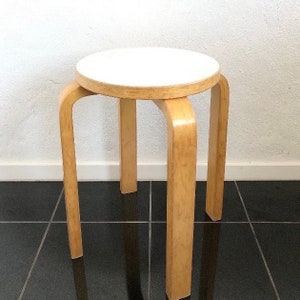 May include: A white and natural wood stool with a round seat and three legs. The stool is on a black tiled floor.