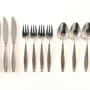 Könnte beinhalten: Ein Set aus Edelstahlbesteck, bestehend aus Messern, Gabeln und Löffeln. Die Messer haben eine gebogene Klinge und einen langen, schlanken Griff. Die Gabeln haben vier Zinken und die Löffel eine ovale Schale. Das Besteck ist auf einer weißen Oberfläche angeordnet.