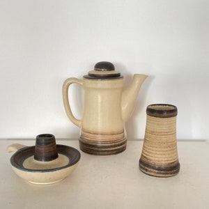 May include: A ceramic coffee pot with a lid, a small candle holder, and a vase. The items are beige with brown horizontal stripes and accents. The coffee pot has a handle and spout. The candle holder has a circular base and a central cylinder. The vase is cylindrical.