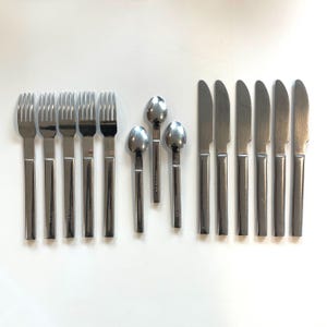 May include: A set of stainless steel flatware, including forks, spoons, and knives, arranged on a white surface. The utensils have a simple, modern design with rectangular handles and a polished finish. The set includes six forks, three spoons, and six knives.