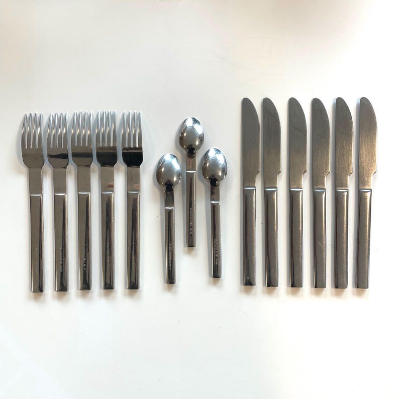 May include: A set of stainless steel flatware, including forks, spoons, and knives, arranged on a white surface. The utensils have a simple, modern design with rectangular handles and a polished finish. The set includes six forks, three spoons, and six knives.