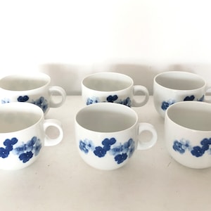 Könnte beinhalten: Sechs weiße Keramik-Kaffeetassen mit blauen Blumenmustern. Die Tassen haben eine abgerundete Form und einen Henkel auf der rechten Seite.