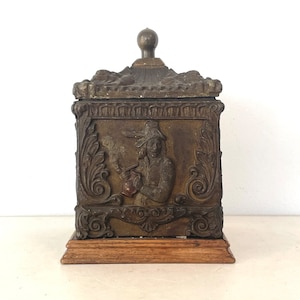 Puede incluir: Caja antigua de color bronce con tapa y base de madera. La caja presenta un relieve detallado de una persona fumando en pipa, rodeada de adornos. La tapa está rematada con un pequeño pomo redondo.