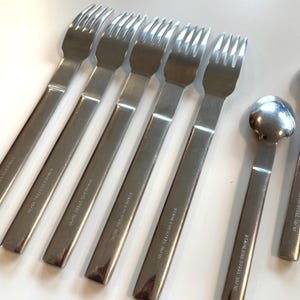 May include: A set of stainless steel forks and spoons. The forks have four tines and long, rectangular handles. The spoons have a rounded bowl. The text "STAINLESS STEEL 18/10" is printed on the handles.
