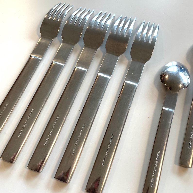 May include: A set of stainless steel forks and spoons. The forks have four tines and long, rectangular handles. The spoons have a rounded bowl. The text "STAINLESS STEEL 18/10" is printed on the handles.