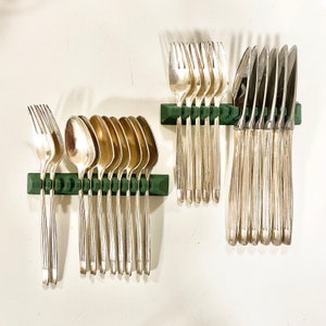 Puede incluir: Un conjunto de cubiertos plateados, que incluye tenedores, cucharas y cuchillos, cuidadosamente dispuestos en soportes verdes. Los utensilios tienen un diseño elegante. La disposición sugiere un entorno de comedor formal.