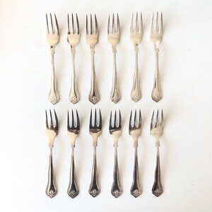 May include: Twelve silver forks arranged on a white surface. Each fork has three tines and a decorative handle with a floral design. The forks are evenly spaced and appear to be vintage or antique.