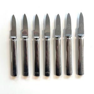 Könnte beinhalten: Set aus sieben silbernen Buttermessern. Jedes Messer hat eine spitze Klinge und einen langen, zylindrischen Griff. Die Griffe haben eine leichte Verjüngung und ein dekoratives Emblem in der Nähe der Klinge. Die Messer sind in einer Reihe auf einer weißen Oberfläche angeordnet.