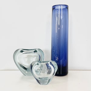 Può includere: Tre vasi di vetro, uno alto e cilindrico con un gradiente di colore blu, e due più piccoli a forma di cuore in vetro trasparente.