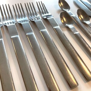 May include: A collection of stainless steel silverware, including forks and spoons, arranged on a light-colored surface. The silverware has a simple, modern design with long, rectangular handles and a polished finish. The forks have four tines.