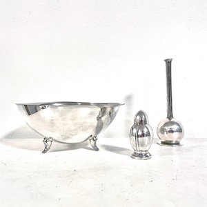 Puede incluir: Colección de tres objetos plateados: un cuenco grande con tres patas, un pequeño salero y un jarrón alto con base redonda. Los objetos están dispuestos sobre una superficie blanca.