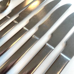 May include: Close-up of a set of stainless steel dinner knives. The knives have a simple, modern design with a rounded blade and a slightly curved handle. The blades are shiny and reflect the light.