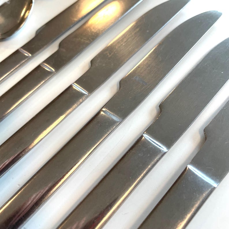 May include: Close-up of a set of stainless steel dinner knives. The knives have a simple, modern design with a rounded blade and a slightly curved handle. The blades are shiny and reflect the light.