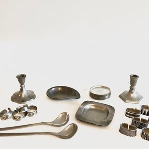 May include: An assortment of vintage silver tableware and decorative items. The collection includes two candlesticks, a small dish, a stack of coasters, a square plate, a set of napkin rings, and serving spoons. The napkin rings feature various geometric designs.