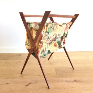 May include: A vintage sewing basket with a wooden frame and a floral fabric bag. The frame is made of dark wood and has a cross-legged design. The fabric bag features a yellow background with pink, red, and green floral patterns.