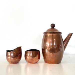 May include: A copper tea set with a teapot, creamer, and sugar bowl. The teapot has a wooden knob on the lid and a spout. The creamer and sugar bowl are small and rounded. The set is displayed on a white surface.