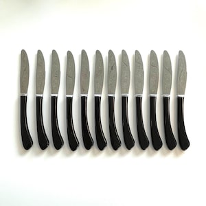 Könnte beinhalten: Set aus zwölf Buttermessern. Jedes Messer hat eine silberne Klinge mit strukturiertem Muster und einen schwarzen, gebogenen Griff. Die Messer sind in einer Reihe vor weißem Hintergrund angeordnet, was ihr elegantes Design hervorhebt.