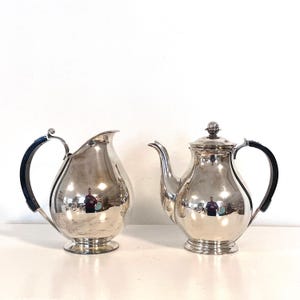 以下が含まれることがあります： 銀色のティーセット。ティーポットとクリーマーが含まれています。ティーポットには蓋と曲がった注ぎ口があります。どちらのアイテムにも黒いハンドルが付いています。セットは白い表面に置かれています。