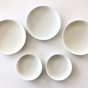 May include: A set of five white ceramic plates. The plates are arranged in a circular pattern, with three larger plates and two smaller plates. The plates have a subtle, textured wood grain pattern.