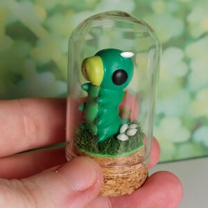 Puede incluir: Una figura de oruga verde y amarilla dentro de una pequeña campana de cristal. La figura se asienta sobre un lecho de césped artificial verde y pequeñas flores blancas, sobre una base de corcho. La campana se sostiene en una mano.