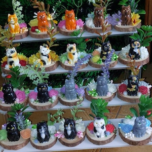 May include: A display of decorative cat figurines in various colors, including orange, black, gray, and brown. Each cat sits on a small wooden slice surrounded by artificial flowers and greenery. The figurines are arranged on tiered shelves.