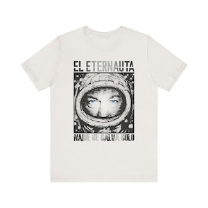 Puede incluir: Camiseta blanca con un gráfico en blanco y negro de una persona con un casco espacial y ojos azules. El texto "EL ETERNAUTA" está encima del gráfico y "NADIE SE SALVA SOLO" debajo.