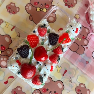 Op de afbeelding: Een transparante telefoonhoesje versierd met witte frosting, rode kersen, aardbeien en bramen. De hoes is versierd met kleine witte Hello Kitty figuren.