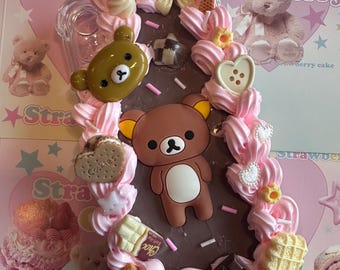 May include: A decorative phone case featuring a brown bear design with pink frosting, bows, and various dessert-themed embellishments. The case includes a clear section for the phone's camera and is surrounded by a pink and brown border.