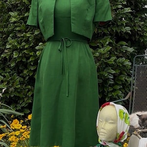 May include: A vintage green dress and matching bolero jacket set. The dress has a tie waist and a knee-length skirt. The jacket has short sleeves and a rounded collar. A pearl necklace is on the mannequin. A head mannequin wears a scarf.