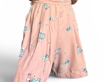 May include: A dusty pink jumpsuit featuring a repeating pattern of figures wearing blue polka-dot skirts and holding white flowers. The garment has a cinched waist with a horizontal band, pleated front details, and wide-cut legs that fall to the mid-thigh.