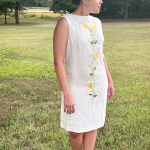 May include: Cream-colored sleeveless dress with a floral design. The dress features yellow flowers and green leaves embroidered down the front. The dress is knee-length and has a simple, elegant design, suitable for a summer day.
