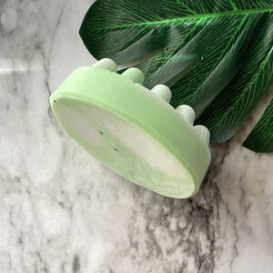 May include: A green and white oval-shaped soap bar with raised bumps on the top. The soap is on a marble surface with a green leaf in the background.
