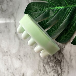 May include: A light green and white silicone facial cleansing brush with five raised bumps. The brush is on a white marble surface with a green leaf in the background.