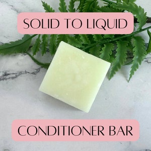 Puede incluir: Una barra de acondicionador blanca con el texto "SOLID TO LIQUID" y "CONDITIONER BAR" sobre una superficie de mármol blanco con follaje verde.