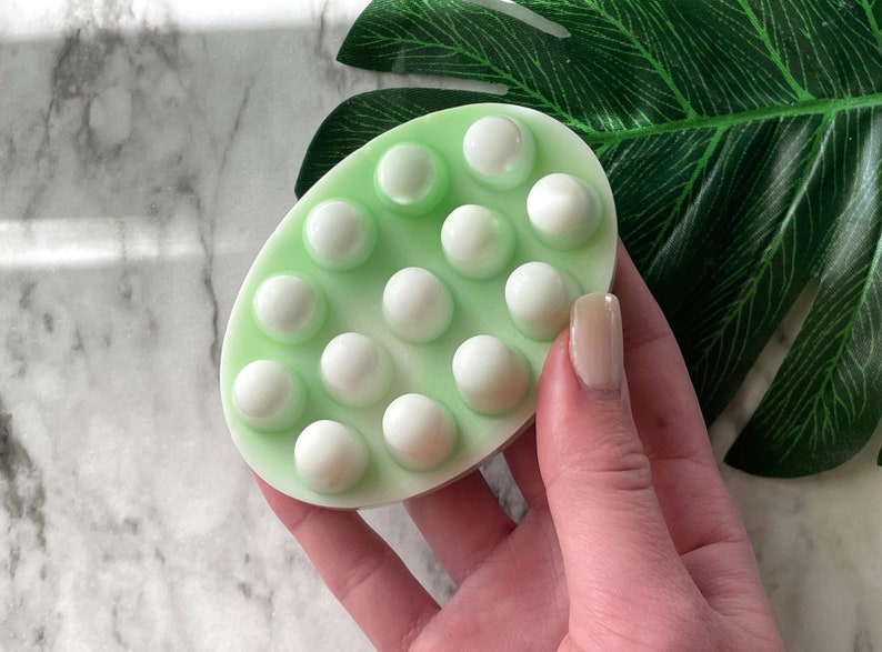 May include: A light green oval-shaped bath scrub with white raised bumps. The scrub is being held in a hand.