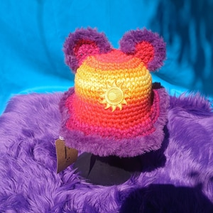 May include: A colorful, handmade crochet hat with a sun emblem. The hat features a gradient of yellow, orange, and pink, with purple faux fur trim and ear-like accents. The hat is displayed on a purple furry surface.