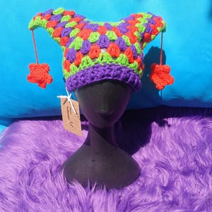 May include: A colorful, handmade crochet jester hat. The hat features a purple brim, a patchwork of red, green, and purple squares, and two pointed tips. Each tip has a dangling red flower. The hat is displayed on a black mannequin head.