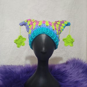 Könnte beinhalten: Ein gehäkelter Hut mit spitzen Ohren und grünen Sternanhängern. Der Hut zeigt eine Mischung aus Blau, Lila und Grün. Er wird auf einem schwarzen Schaufensterkopf vor einem lila, flauschigen Hintergrund präsentiert. Das Design ist detailreich.