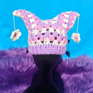 Può includere: Un cappello all'uncinetto fatto a mano dal design stravagante. Il cappello presenta un bordo lilla e un motivo patchwork di quadrati bianchi, rosa e lavanda. Sono attaccati due fiori pendenti. Il cappello è esposto su una testa di manichino nera su sfondo blu.