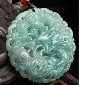 Puede incluir: Un colgante de jade tallado con un diseño detallado de dos dragones entrelazados. El colgante es de color verde claro y tiene un acabado liso y pulido.